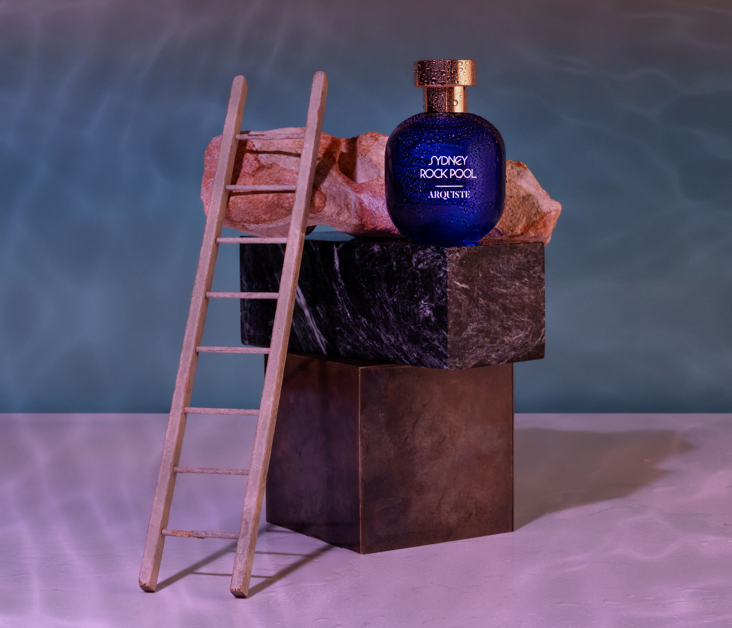 Arquiste Sydney Rock Pool perfume bottle on a rock with a ladder leading up to it against a textured background.
